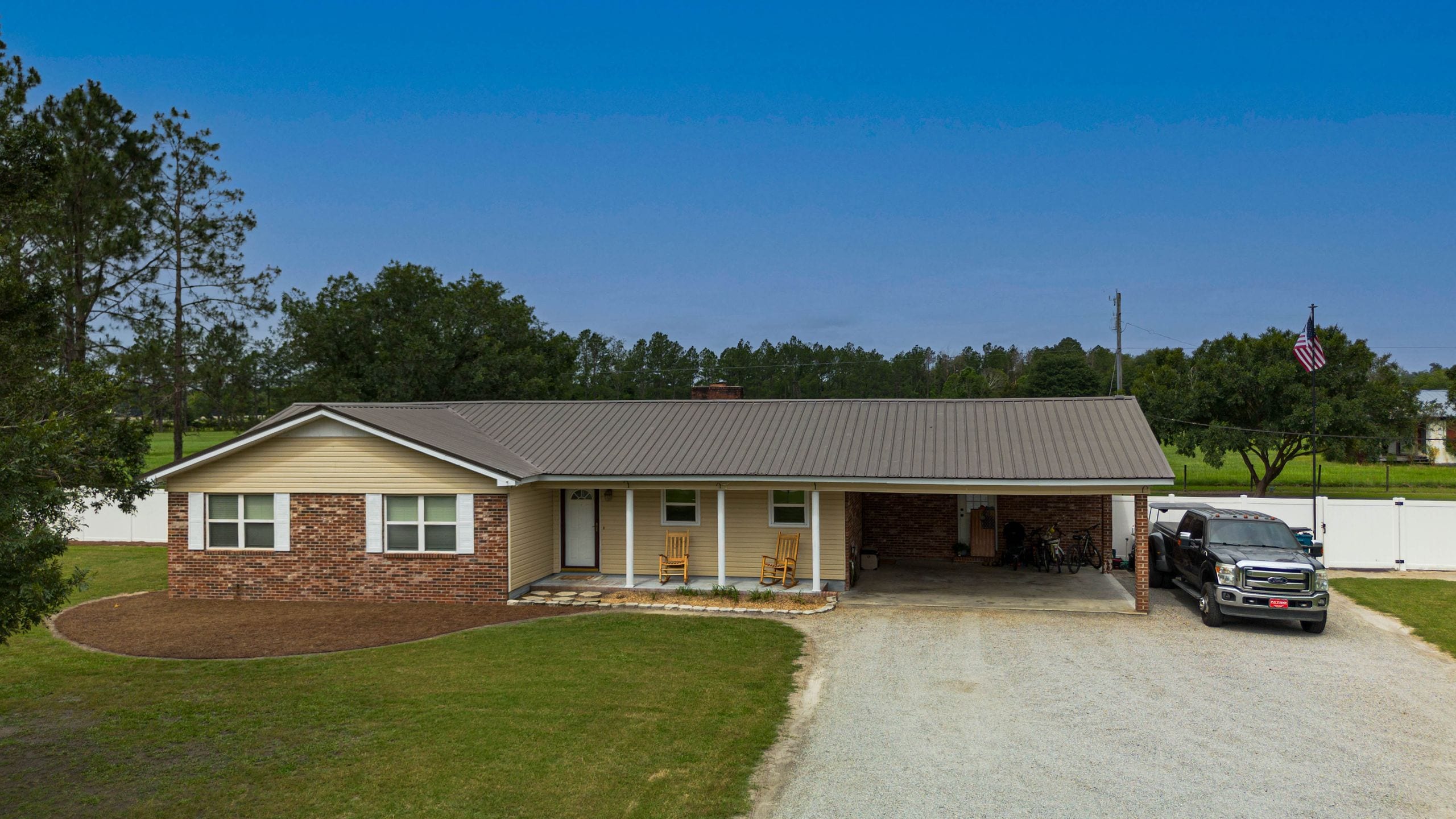 Brick and cream ranch home with a brown metal roof and open carport, twin rocking chairs on the front porch and a pickup truck parked on the gravel drive